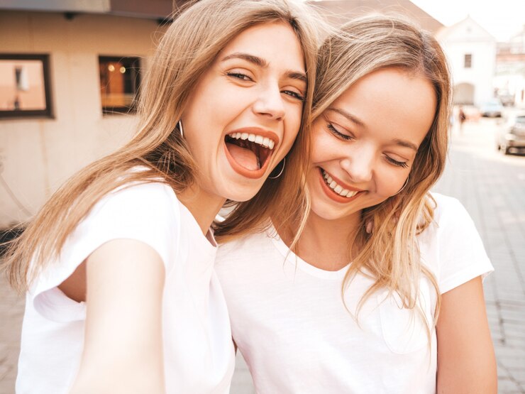 Foto gratuito due giovani donne bionde sorridenti dei pantaloni a vita bassa in vestiti bianchi della maglietta di estate. ragazze che prendono le foto dell'autoritratto del selfie sullo smartphone.
