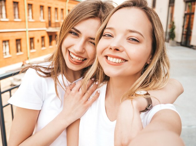 Due giovani donne bionde sorridenti dei pantaloni a vita bassa in vestiti bianchi della maglietta di estate. Ragazze che prendono le foto dell'autoritratto del selfie sullo smartphone.