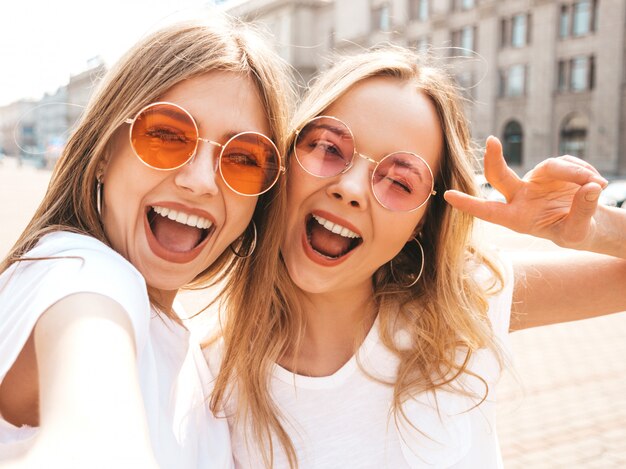 Due giovani donne bionde sorridenti dei pantaloni a vita bassa in vestiti bianchi della maglietta di estate.