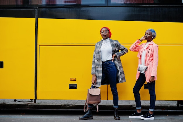 Due giovani donne musulmane africane alte e magre attraenti moderne alla moda in hijab o turbante, sciarpa e cappotto poste contro l'autobus giallo