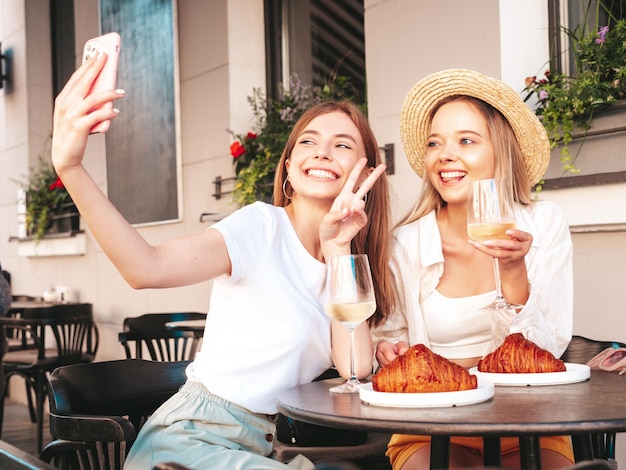 Due giovani e belle donne hipster sorridenti in abiti estivi alla moda Donne spensierate in posa al bar sulla veranda in stradaModelli positivi che bevono vino biancoMangiare croissant Scattare foto selfie