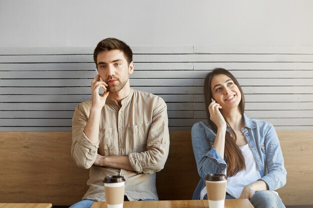 Due giovani in una lite seduti uno accanto a altro nella caffetteria, guardando da parte, bevendo caffè e parlando al telefono.