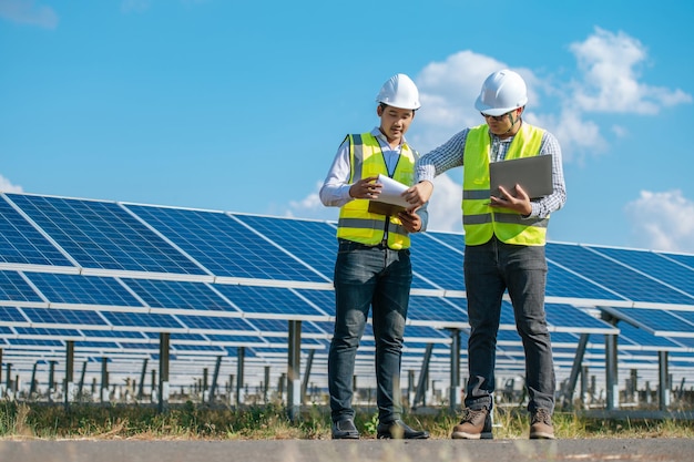 Due giovani ingegneri asiatici che camminano lungo le file di pannelli fotovoltaici nella fattoria solare Usano il computer portatile e parlano insieme