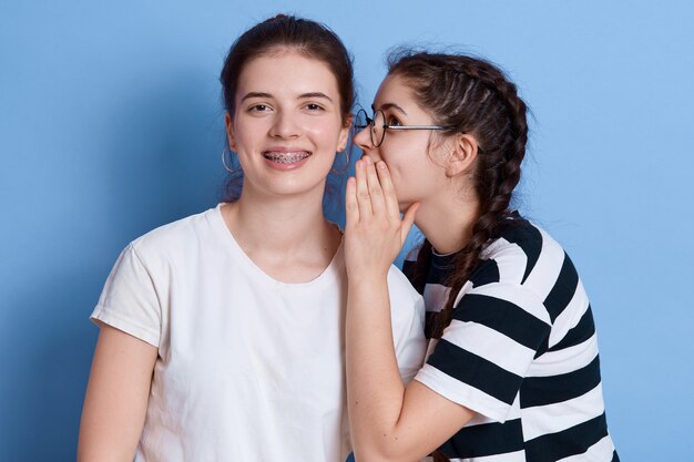Due giovani ragazze eccitate vestite in abiti estivi che pettegolano isolato, signora con gli occhiali che bisbigliano qualcosa all'orecchio, ragazze che esprimono felicità.