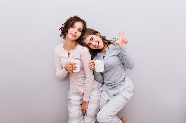 Due giovani ragazze in pigiama con coppe sul muro grigio. Ragazza con capelli lunghi magra testa sulla spalla della ragazza con i capelli ricci. Sorridono.