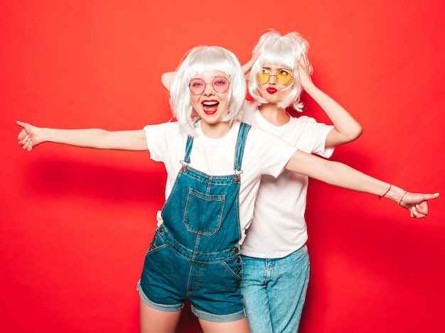 Due giovani ragazze sexy hipster in parrucche bianche e labbra rosse. Belle donne alla moda in abiti estivi. Modelle vivaci in posa vicino al muro rosso in studio estate in occhiali da sole