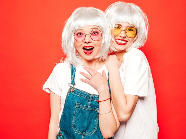 Due giovani ragazze sorridenti sexy dei pantaloni a vita bassa in parrucche bianche e labbra rosse Belle donne d'avanguardia in vestiti di estate Modelli liberi che posano vicino alla parete rossa nell'estate dello studio che impazzisce
