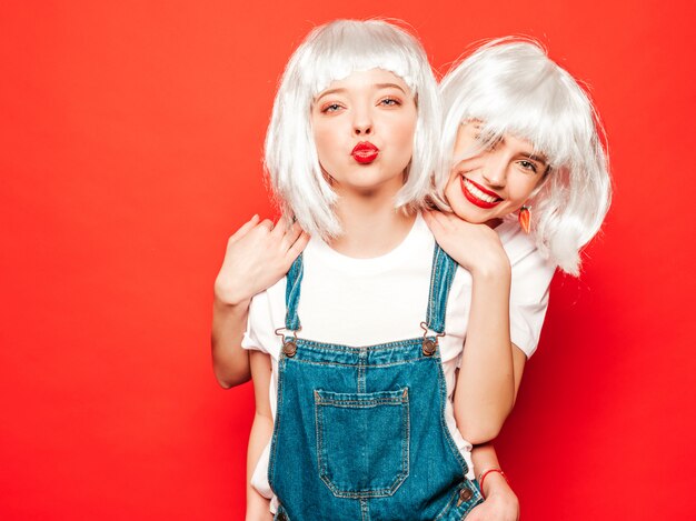 Due giovani ragazze sorridenti sexy dei pantaloni a vita bassa in parrucche bianche e labbra rosse Belle donne d'avanguardia in vestiti di estate Modelli liberi che posano vicino alla parete rossa nell'estate dello studio che impazzisce