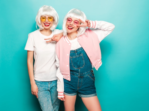 Due giovani ragazze sorridenti sexy dei pantaloni a vita bassa in parrucche e labbra rosse Belle donne d'avanguardia in vestiti di estate Modelli liberi che posano vicino alla parete blu in estate dello studio mostra la lingua e il segno di pace