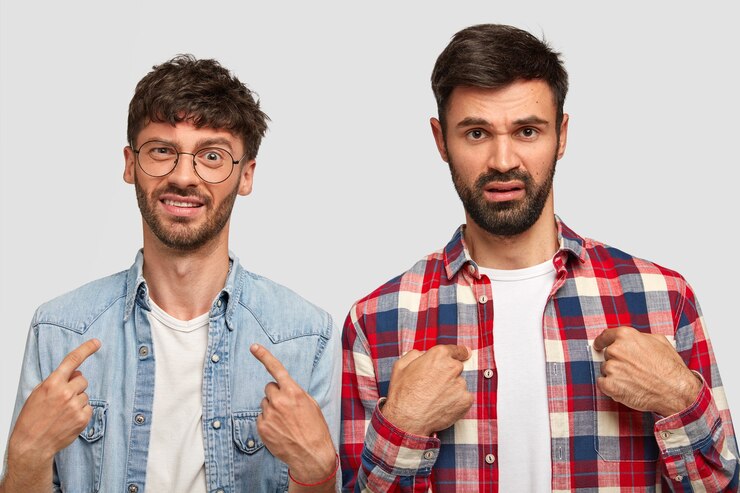 Foto gratuito due giovani scontenti si indicano, chiedono perché dovrebbero fare i doveri per la casa, aggrottano le sopracciglia per lo scontento, indossano camicie eleganti, hanno stoppie scure, isolato su un muro bianco
