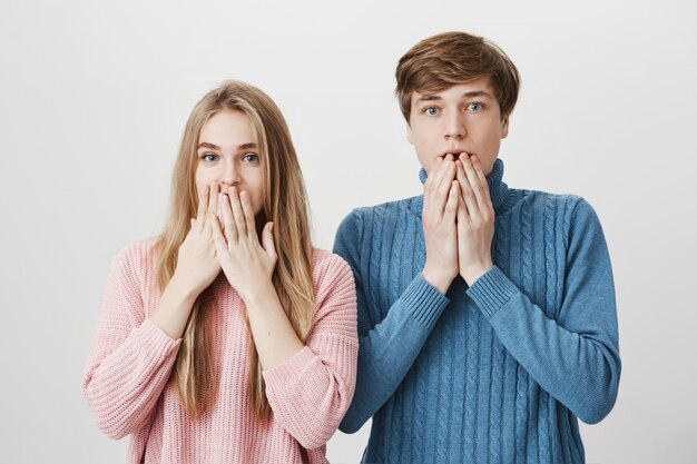 Due giovani studenti inorriditi e stupiti in maglioni eleganti che fissano la telecamera increduli, tenendo la bocca aperta,