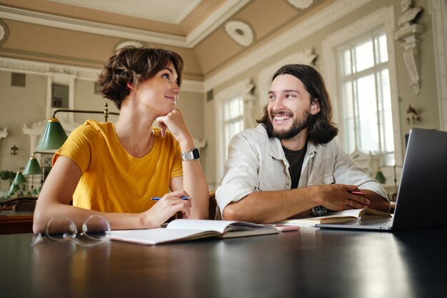 Due giovani studenti sorridenti che lavorano felicemente su un progetto di studio con laptop e libri nella biblioteca dell'università