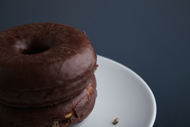 Due gustose ciambelle appena sfornate glassate al cioccolato su un piccolo piatto in ceramica bianca isolato nell'angolo del vecchio tavolo di legno blu rustico. Messa a fuoco ravvicinata con dettagli