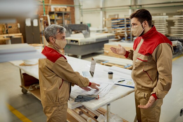 Due lavoratori con maschere facciali che discutono dei piani di progetto in un'officina di falegnameria