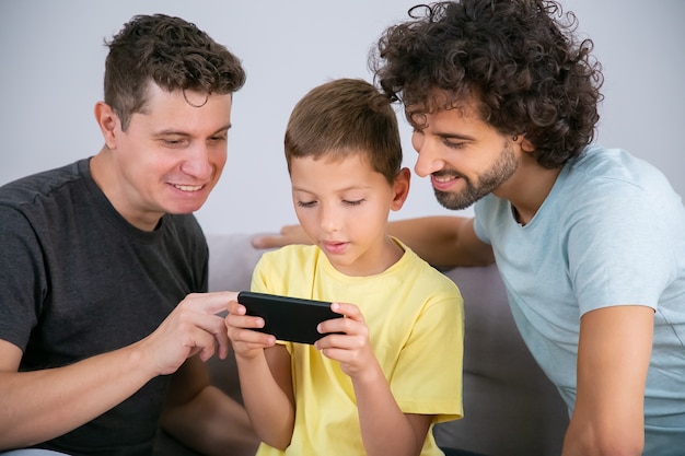 Due papà felici che insegnano al figlio a usare l'app online sul cellulare. Ragazzo che gioca gioco sul telefono cellulare. Famiglia a casa e concetto di comunicazione