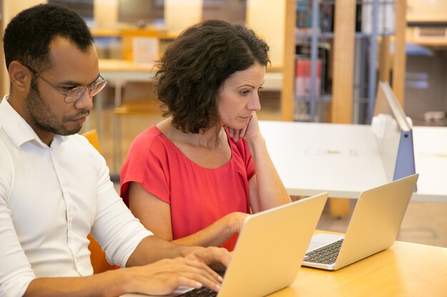 Due persone riflessive, seduto con i computer portatili in biblioteca