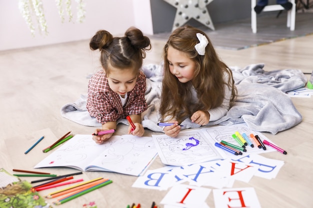 Due piccole ragazze carine che stanno disegnando nel libro da colorare che giace sul pavimento sulla coperta e imparano le lettere