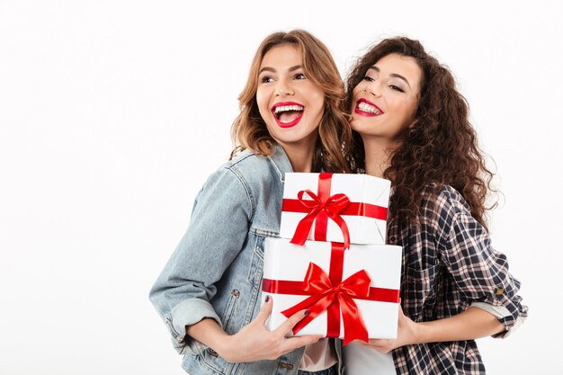 Due ragazze allegre che posano con i regali e che distolgono lo sguardo sopra la parete bianca