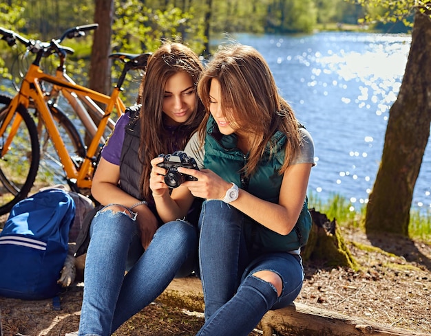 Due ragazze brune carine che utilizzano una fotocamera compatta e si rilassano dopo il giro in bicicletta sulla selvaggia costa del fiume.