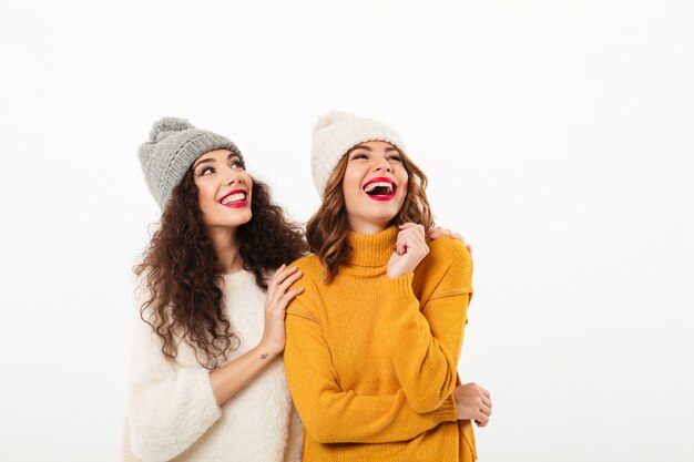 Due ragazze che ridono in maglioni e cappelli che stanno insieme mentre guardano su sopra il muro bianco