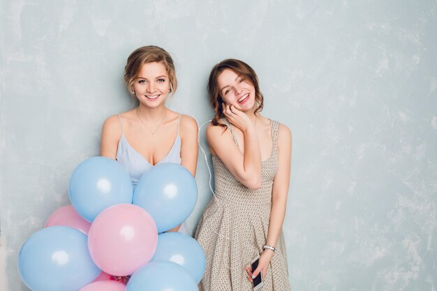 Due ragazze che si divertono in uno studio e giocano con palloncini blu e rosa. La ragazza bionda indossa un vestito blu. La ragazza bruna indossa un vestito leggero e tiene lo smartphone ascoltando musica sugli auricolari.