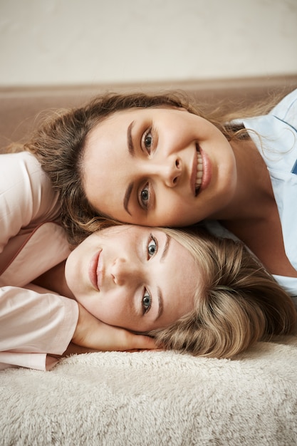 Due ragazze con una relazione stretta, sentendosi come sorelle. Colpo verticale di belle donne che si trovano sul sofà e che sorridono ampiamente. Affascinante amica dai capelli ricci che giace sulla testa del suo bestie