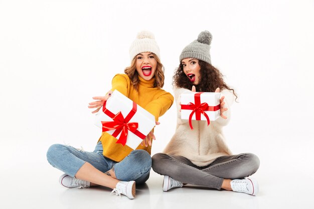 Due ragazze di grido felici in maglioni e cappelli che si siedono insieme con i regali sul pavimento sopra la parete bianca