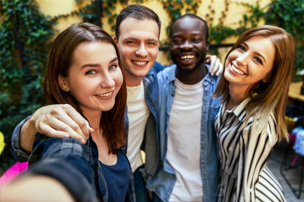 Due ragazze e due ragazzi stanno scattando selfie all'aperto, si abbracciano e con sorrisi sinceri