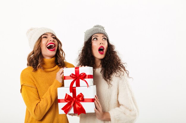 Due ragazze felici sorprese in maglioni e cappelli che stanno insieme mentre tengono i regali e guardano su sopra la parete bianca