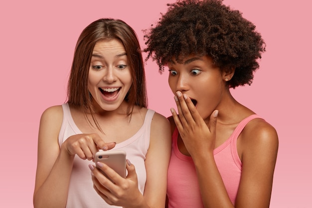 Due ragazze multietniche guardano il fantastico articolo sulla pagina web tramite cellulare, hanno sorpreso espressioni felici, vestite con abiti estivi, usano internet wireless, modella contro il muro rosa