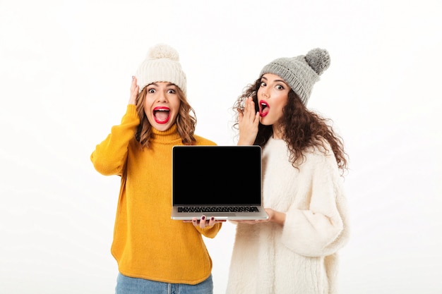 Due ragazze scioccate in maglioni e cappelli in piedi insieme mentre mostra lo schermo del computer portatile in bianco sul muro bianco