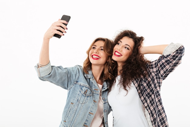 Due ragazze sorridenti che posano insieme e che fanno selfie sullo smartphone sopra la parete bianca