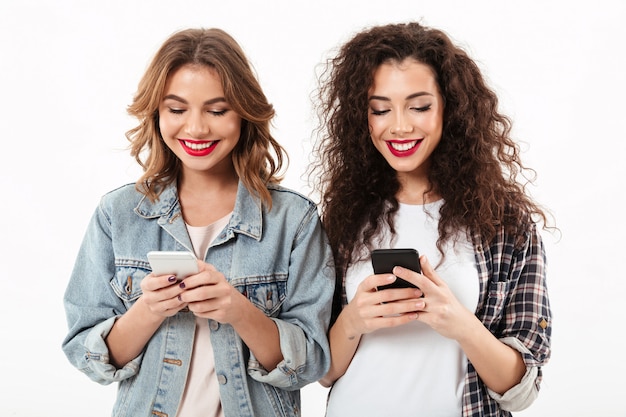 due ragazze sorridenti che scrivono messaggio sullo smartphone sopra la parete bianca