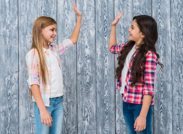 Due ragazze sorridenti che stanno contro la parete di legno grigia che dà livello cinque