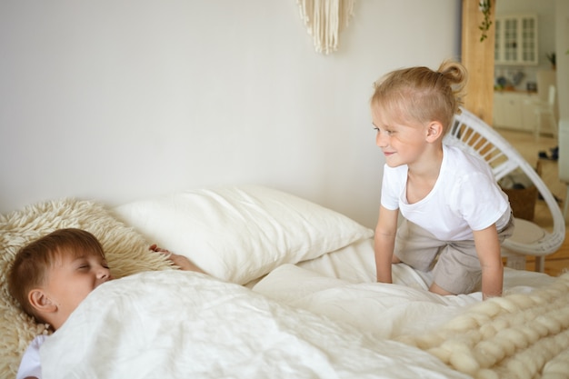 Due ragazzi che giocano a letto. Ragazzino biondo sveglio che si siede sui vestiti da letto bianchi guardando il suo fratello anziano che finge di dormire. Bambini che giocano in camera da letto. Famiglia, infanzia e divertimento