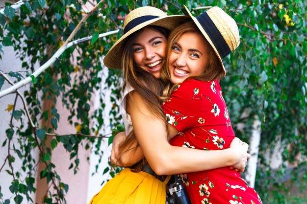Due sorelle abbastanza giovani felici, abbracci sorridenti ridendo e divertendosi insieme, portando vestiti e cappelli femminili vintage retrò alla moda. All'aperto.
