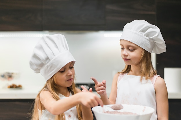 Due sorelle in cucina indossando il cappello da cuoco guardando il dito disordinato mentre si prende la polvere di cacao