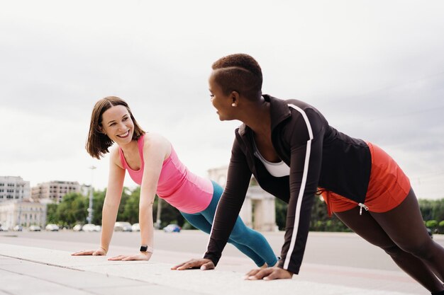 Due sorridenti diverse giovani donne in abiti da allenamento atletico stanno facendo un esercizio Plank lavorando insieme nella piazza della città