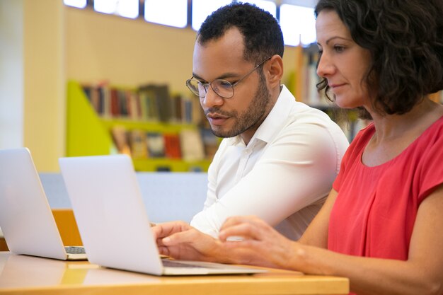 Due studenti concentrati che parlano e che esaminano computer portatile la biblioteca