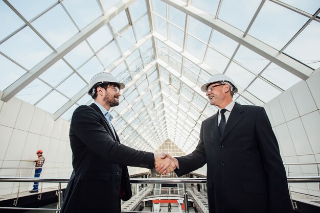 Due uomini d'affari allegri che si stringono la mano nel casco al nuovo edificio