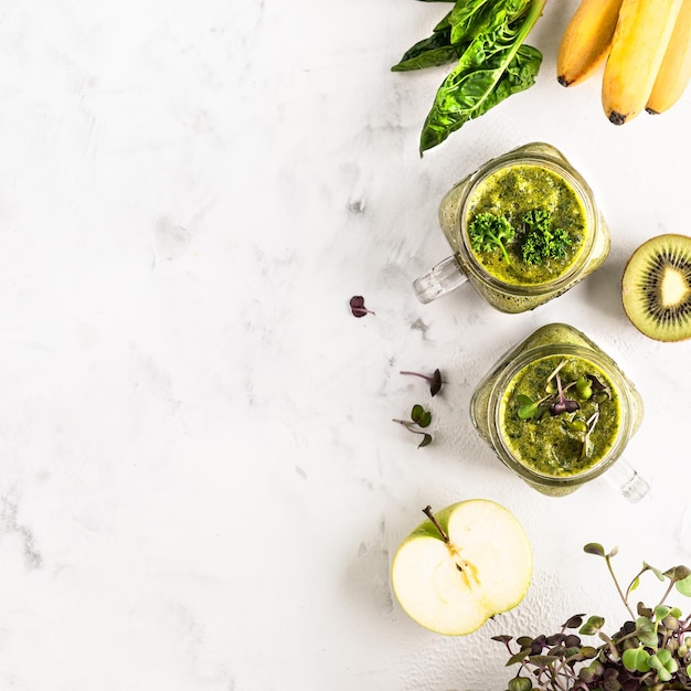 Due vasetti di vetro di cocktail verde fatto in casa a base di spinaci con mela banana e kiwi e verdure bagnate su sfondo bianco vista dall'alto