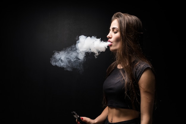 E - sigaretta vaping della giovane bella donna sulla parete nera