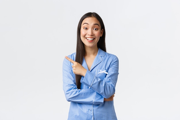 Eccitata bella ragazza asiatica in pigiama blu che punta il dito a sinistra e sorride entusiasta, mostrando un fantastico banner promozionale, raccontando il prodotto, facendo annuncio su sfondo bianco.