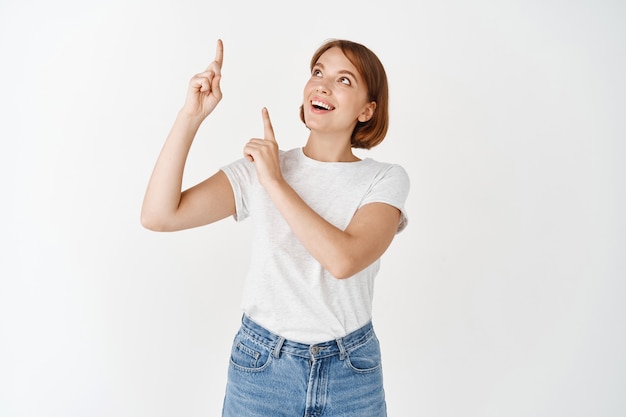 Eccitata bella signora in t-shirt che punta le dita in alto, sorride e sembra divertita, controlla la pubblicità promozionale, in piedi sul muro bianco