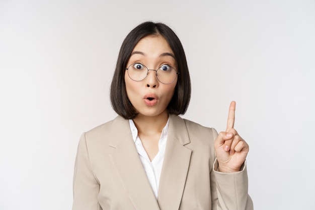 Eccitata donna asiatica con gli occhiali alzando il dito segno eureka ha un'idea in piedi su sfondo bianco Copia spazio