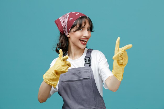 Eccitata giovane donna addetta alle pulizie che indossa bandana uniforme e guanti di gomma guardando la fotocamera che ti mostra il gesto isolato su sfondo blu