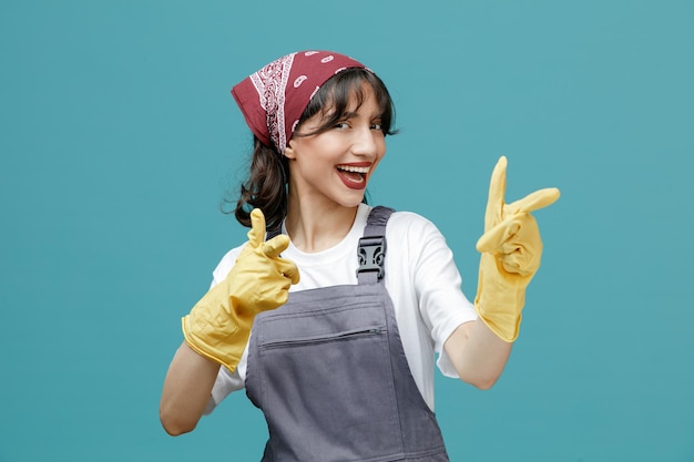 Eccitata giovane donna addetta alle pulizie che indossa bandana uniforme e guanti di gomma guardando la fotocamera che ti mostra il gesto isolato su sfondo blu