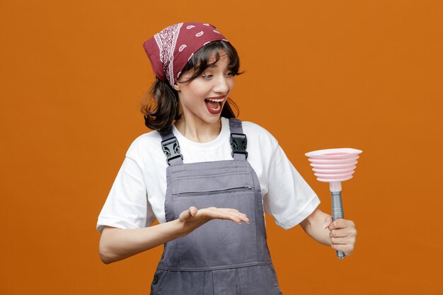 Eccitata giovane donna addetta alle pulizie che indossa uniforme e bandana tenendo lo stantuffo guardandolo mostrando la mano vuota isolata su sfondo arancione