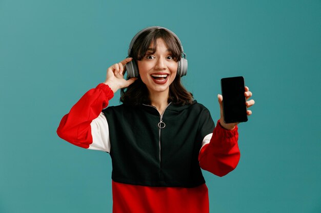 Eccitata giovane donna caucasica che indossa e afferra le cuffie guardando la fotocamera che mostra il telefono cellulare isolato su sfondo blu