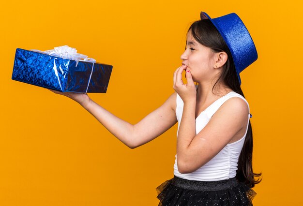 Eccitata giovane ragazza caucasica con cappello da festa blu che tiene e guarda la confezione regalo mettendo la mano sulla bocca isolata sulla parete arancione con spazio di copia